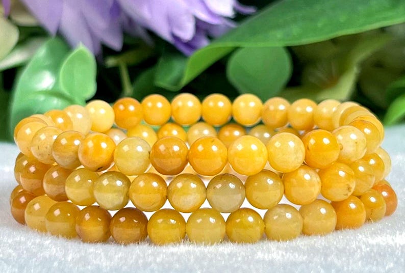 May include: Three bracelets of round, yellow-orange beads. The beads have a smooth, polished appearance and vary slightly in color. The bracelets are stacked, with a blurred background of green leaves and purple flowers.