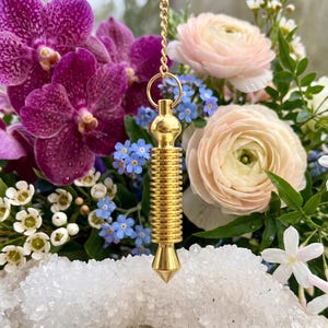May include: A gold-toned metal pendulum with a ribbed, cylindrical body and a pointed tip, suspended from a delicate gold-colored chain. The pendulum is positioned in front of a vibrant floral arrangement featuring purple orchids and pale pink ranunculus.