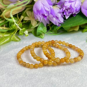 May include: Three beaded bracelets with golden-yellow and orange beads. The bracelets are arranged on a white surface, with purple flowers and green foliage in the background. The beads appear to be approximately 0.3 inches in diameter.