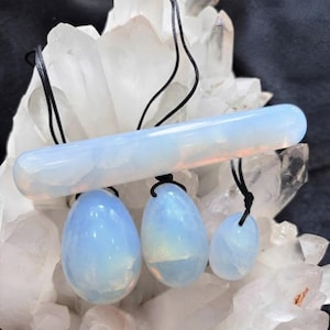 Puede incluir: Un conjunto de cuatro piedras de opalita translúcidas de color blanco lechoso, que consta de un cilindro horizontal y tres piezas en forma de huevo de diferentes tamaños. Cada piedra está suspendida por un fino cordón negro.