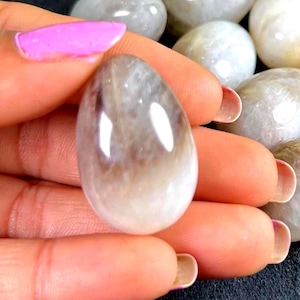 May include: A polished, oval-shaped gemstone with a gradient of colors, from a light, creamy white at the base to a darker, brownish-gray at the top. The stone has a smooth, reflective surface, and is held in a hand with pink nail polish.