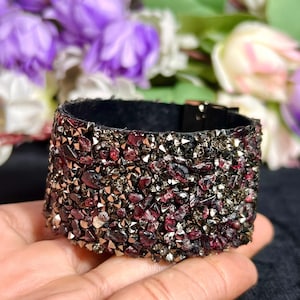 May include: A wide cuff bracelet featuring a dense surface of small, faceted dark red and metallic grey stones. The band has a black interior lining and a black metal buckle closure, creating a textured, multi-dimensional appearance.