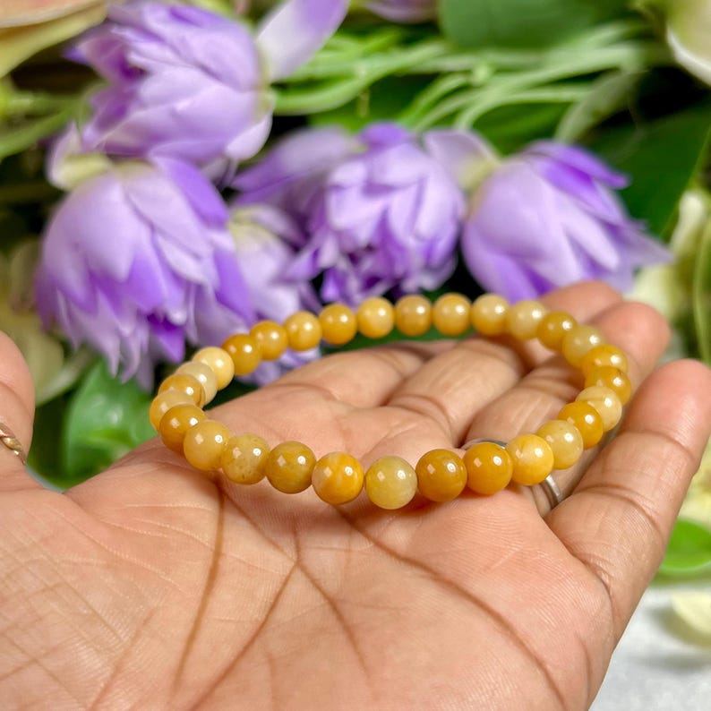 May include: A golden yellow beaded bracelet. The bracelet is made of small, round beads and is displayed against a background of purple flowers and green foliage. The bracelet is held in a hand.