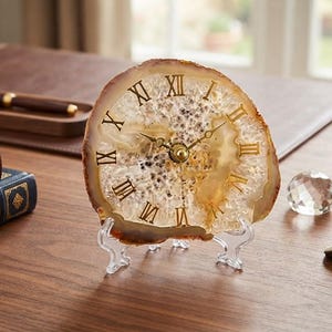 May include: A table clock crafted from a circular slice of translucent beige and white agate stone. The clock face features gold-colored Roman numerals and thin gold hands, supported by a clear acrylic stand on a wooden surface.