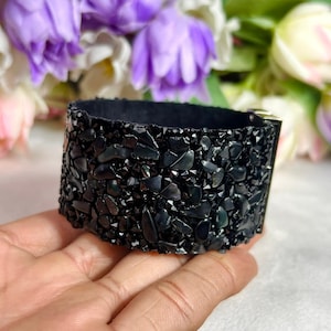 May include: A wide cuff bracelet featuring a dense surface of small, irregular, glossy black stones. The bracelet has a dark interior lining and a metallic silver-toned clasp mechanism visible on the side, creating a textured, dark appearance.