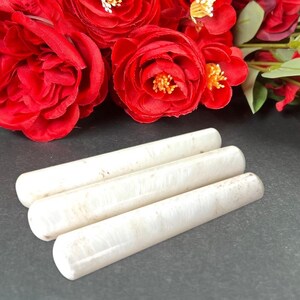 Puede incluir: Tres objetos cil&iacute;ndricos blanquecinos se disponen sobre una superficie oscura, con rosas rojas brillantes de fondo. Los objetos tienen un aspecto liso y ligeramente texturizado. El conjunto es sencillo y elegante.