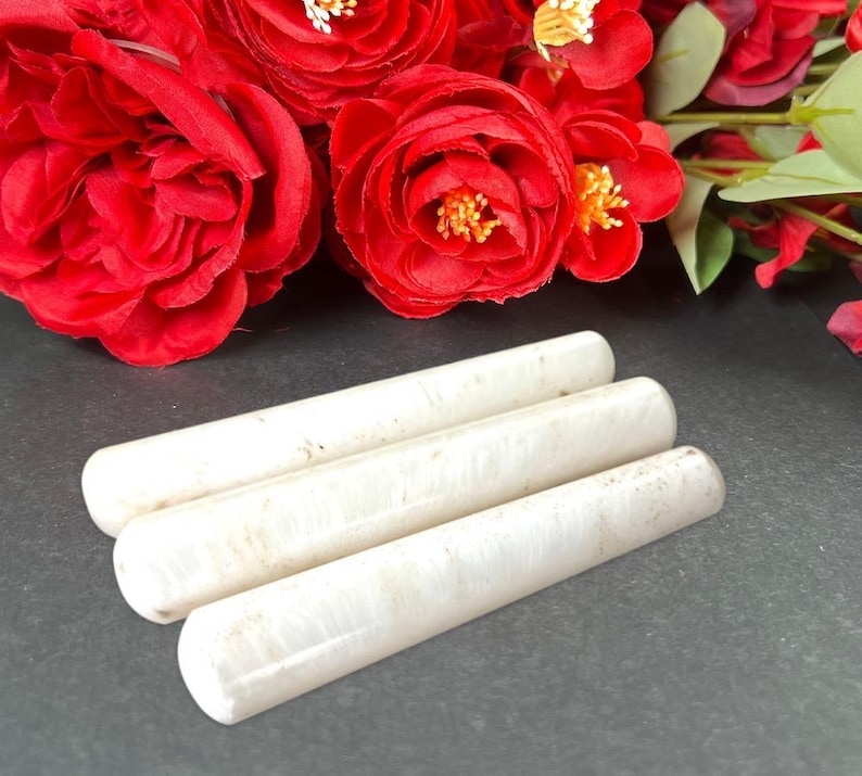 Puede incluir: Tres objetos cil&iacute;ndricos blanquecinos se disponen sobre una superficie oscura, con rosas rojas brillantes de fondo. Los objetos tienen un aspecto liso y ligeramente texturizado. El conjunto es sencillo y elegante.