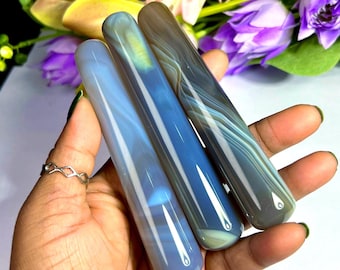 May include: Three polished, cylindrical stones in shades of blue and gray are held in a hand. The stones have a smooth, glossy finish with natural banding patterns. The background features colorful flowers.