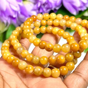 May include: Three strands of yellow-orange beaded bracelets are held in a hand. The beads are round and have a polished finish. The background includes purple flowers and green foliage.