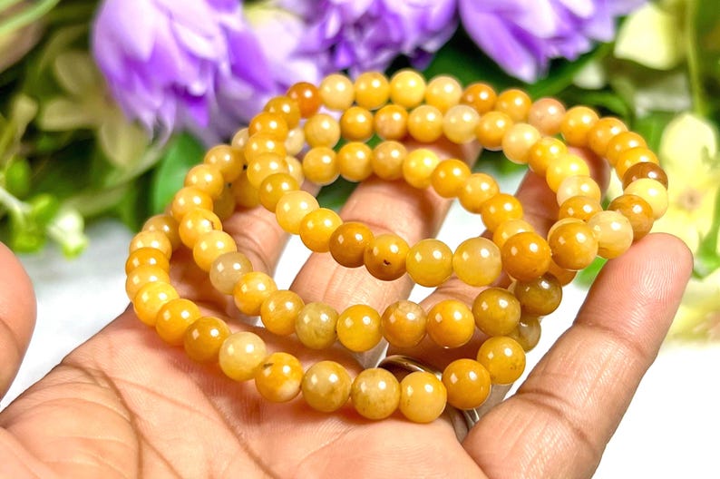May include: Three strands of yellow-orange beaded bracelets are held in a hand. The beads are round and have a polished finish. The background includes purple flowers and green foliage.