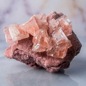 May include: A raw mineral specimen featuring translucent, pale pink cubic crystals clustered atop a textured, dusty rose-coloured rock base. The crystalline structures exhibit sharp geometric edges and internal light refraction, contrasting with the matte, granular surface of the underlying stone formation.