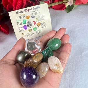 May include: A hand holds a collection of polished crystals, including amethyst, citrine, and malachite, with a card labeled "Money Magnet Crystals." The card lists crystal properties. Red flowers are in the background.