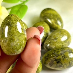 May include: A hand holds a polished, egg-shaped stone with a speckled green and yellow pattern. Other similar stones are in the background. The stones have a glossy finish and are likely used for decorative or spiritual purposes.