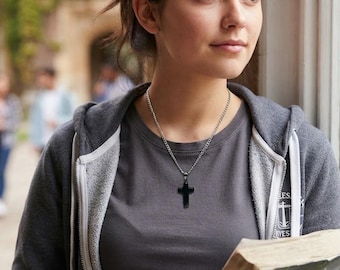 May include: A black cross pendant hangs from a silver-toned metal chain necklace. The cross has a smooth, solid black finish and is suspended from a simple link chain that rests against a grey t-shirt worn under a grey zip-up hooded sweatshirt.