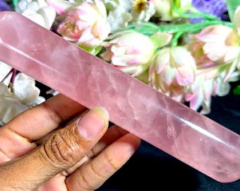 May include: A smooth, cylindrical rose quartz wand with a polished, translucent light pink finish. The stone features natural internal inclusions and a rounded tip, showcasing the crystalline structure of the mineral in a long, slender form.