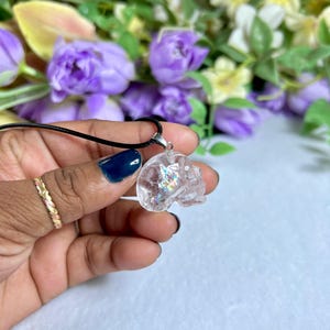 May include: A clear crystal skull pendant on a black cord necklace. The skull is held in a hand with a gold ring and blue nail polish. The background features purple and yellow flowers.