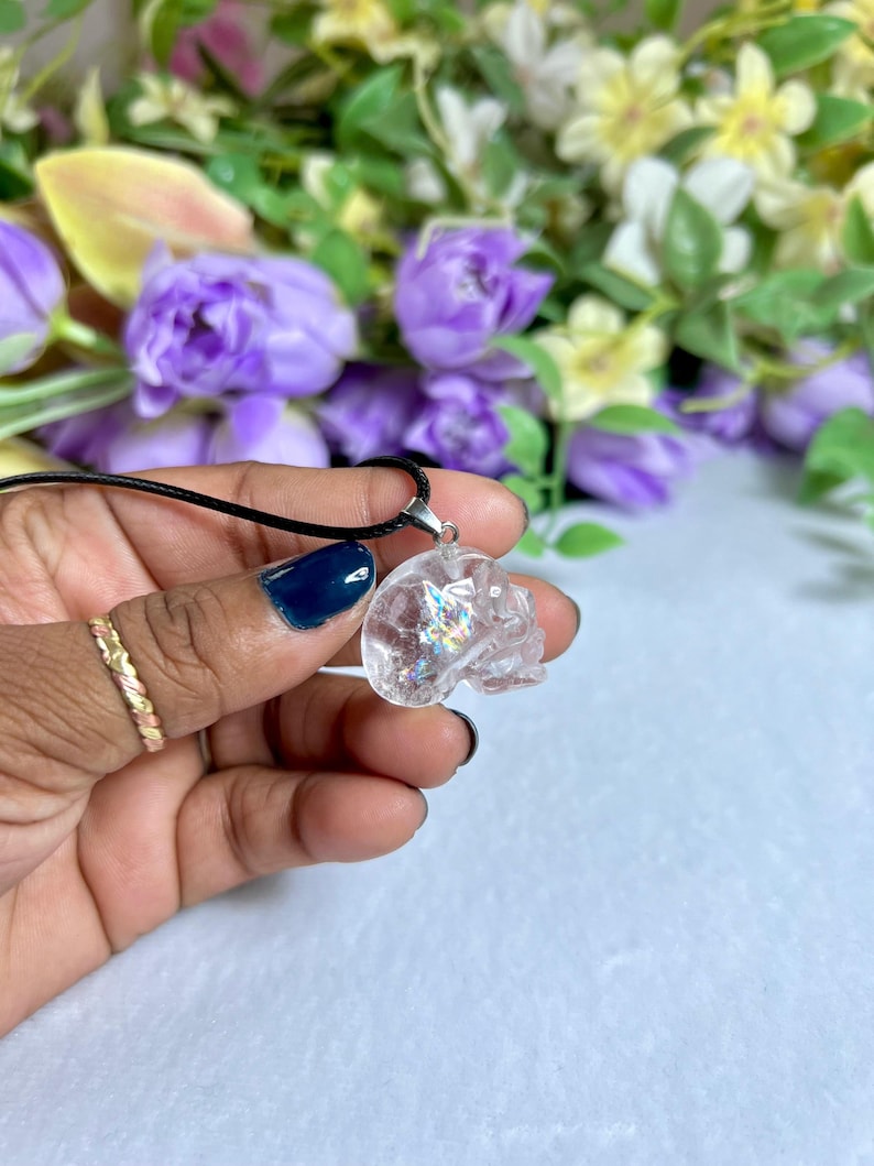 May include: A clear crystal skull pendant on a black cord necklace. The skull is held in a hand with a gold ring and blue nail polish. The background features purple and yellow flowers.