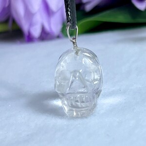 May include: Clear quartz crystal skull pendant hanging from a black cord. The skull is intricately carved with visible facial features. The background features blurred purple flowers and a white surface.