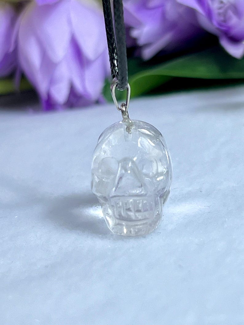May include: Clear quartz crystal skull pendant hanging from a black cord. The skull is intricately carved with visible facial features. The background features blurred purple flowers and a white surface.
