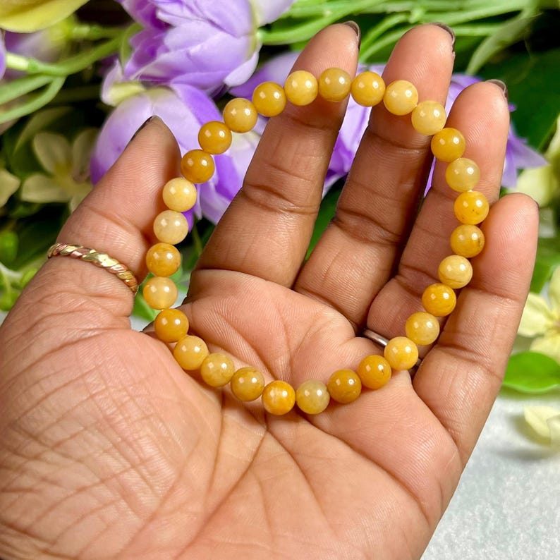 May include: A golden yellow beaded bracelet. The bracelet is made of round, polished beads in varying shades of yellow. The bracelet is held in a hand, with a gold ring visible on the ring finger. The background features purple and yellow flowers.