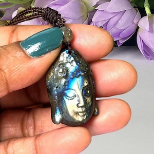 May include: A carved labradorite pendant shaped like a Buddha head, featuring iridescent blue and gold flashes across its surface. The pendant is suspended from a dark brown braided cord, which includes a small, round, polished stone bead.