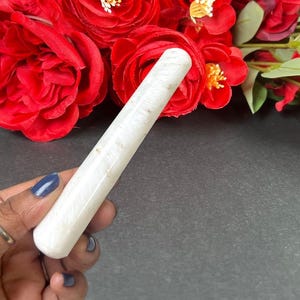 Puede incluir: Una varita de cristal cil&iacute;ndrica blanca con una superficie lisa. La varita se sostiene en una mano con una u&ntilde;a pintada de azul. Rosas rojas en el fondo.