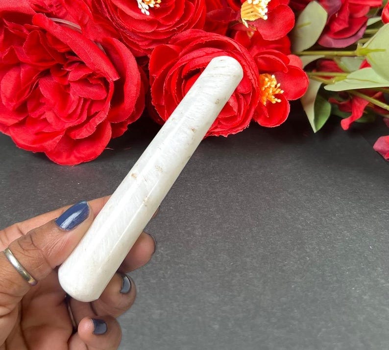 Puede incluir: Una varita de cristal cil&iacute;ndrica blanca con una superficie lisa. La varita se sostiene en una mano con una u&ntilde;a pintada de azul. Rosas rojas en el fondo.