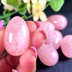 Pode incluir: Uma coleção de ovos de quartzo rosa polidos em vários tamanhos. Os ovos são de uma cor rosa suave e têm uma superfície lisa e brilhante. Alguns ovos estão em foco, enquanto outros estão desfocados no fundo. O fundo inclui folhas verdes e flores amarelas.