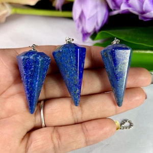 Op de afbeelding: Drie gefacetteerde, blauwe lapis lazuli pendels met zilverkleurige metalen doppen en lussen. De kegelvormige pendels hebben een gepolijste afwerking. De achtergrond bevat paarse bloemen en groene bladeren.