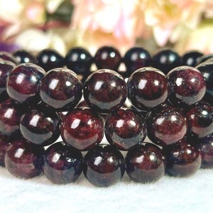 May include: A triple-strand bracelet composed of polished, spherical dark red garnet beads. The beads feature deep burgundy tones with subtle natural inclusions and a reflective, glossy surface finish, arranged in three parallel rows on an elastic cord.