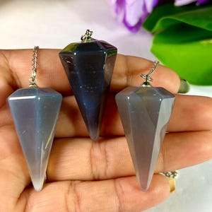 May include: Three faceted crystal pendulums in shades of gray and blue. Each pendulum has a silver chain and a pointed bottom. The crystals are held in a hand, with a blurred background of purple flowers and green leaves.