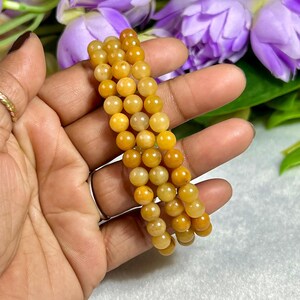 May include: Three strands of yellow-orange gemstone beads are held in a hand. The beads are round and have a polished finish. The background includes purple flowers and green leaves.