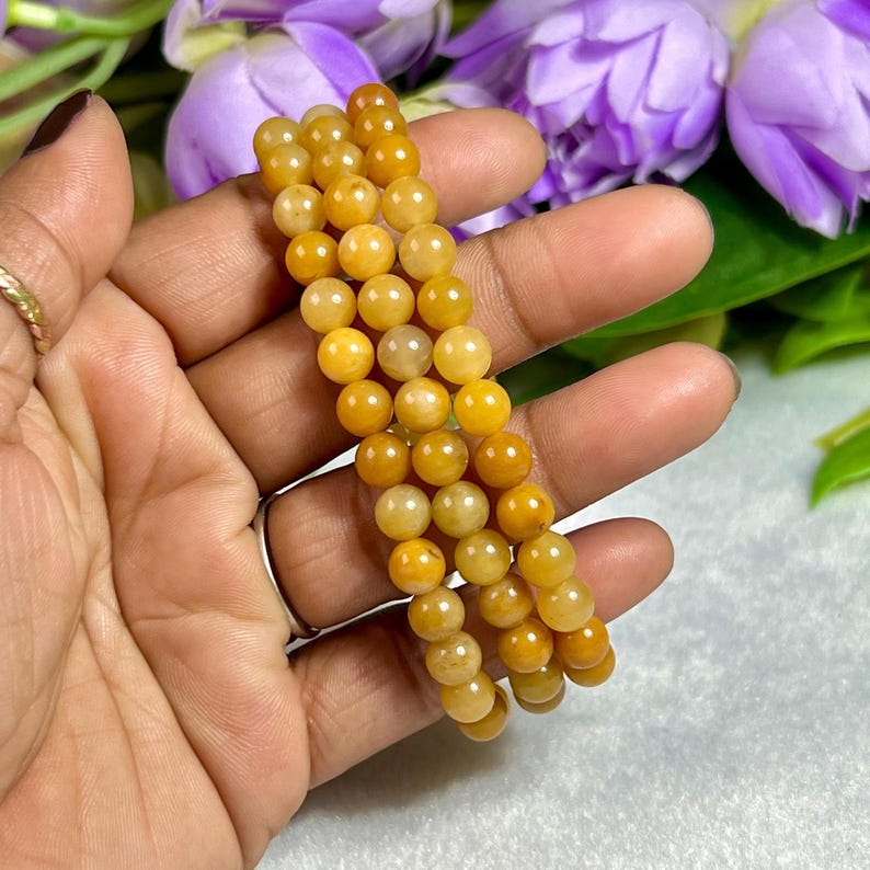 May include: Three strands of yellow-orange gemstone beads are held in a hand. The beads are round and have a polished finish. The background includes purple flowers and green leaves.
