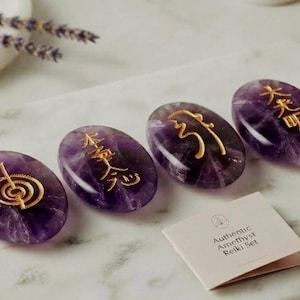 May include: Four oval-shaped, polished purple amethyst stones, each featuring a unique gold-colored engraved symbol. A small, square, off-white card is included, which displays the text "Authentic Amethyst Reiki Set" in black lettering.
