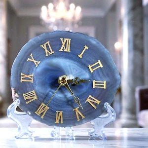 May include: A circular blue agate slice clock featuring gold-colored Roman numerals and matching gold-colored clock hands. The clock is supported by a clear, transparent plastic display stand, showcasing the natural banded patterns of the stone.