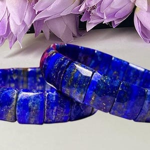 May include: Two stretch bracelets composed of rectangular, faceted lapis lazuli beads. The beads feature a deep royal blue color with natural gold-colored pyrite inclusions and subtle white veining throughout the polished stone surfaces.