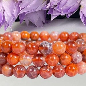 Peut inclure: Un bracelet multi-rangs composé de perles rondes et polies dans des tons variés d'orange, de rouge et de cramoisi profond. Chaque perle présente une texture craquelée distincte et complexe sur toute sa surface, offrant un aspect cohérent.