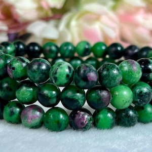 May include: A triple-strand beaded bracelet featuring polished, spherical stones in shades of deep forest green, black, and vibrant magenta. The beads are strung together in a uniform pattern, creating a layered look with a smooth, glossy finish.