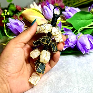 May include: A black and gold wire-wrapped crystal cluster with white and black stones. The cluster has a black loop at the top and is held in a hand. Purple flowers are in the background.