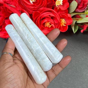 Puede incluir: Tres varitas cil&iacute;ndricas blancas con apariencia marmolada se sostienen en una mano, sobre un fondo de vibrantes rosas rojas. Las varitas tienen extremos redondeados y una textura suave, lo que sugiere que est&aacute;n hechas de piedra o un material similar.