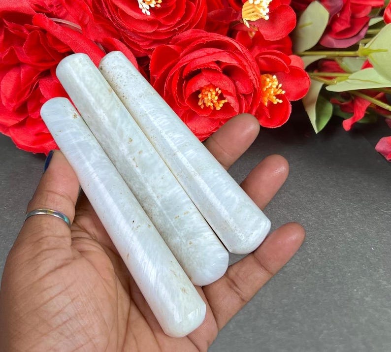 Puede incluir: Tres varitas cil&iacute;ndricas blancas con apariencia marmolada se sostienen en una mano, sobre un fondo de vibrantes rosas rojas. Las varitas tienen extremos redondeados y una textura suave, lo que sugiere que est&aacute;n hechas de piedra o un material similar.