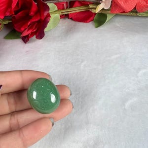 May include: A smooth, oval-shaped green gemstone, held in a hand, with a polished surface. The stone has a speckled appearance and reflects light. Red flowers and greenery are visible in the background, set against a white, textured surface.