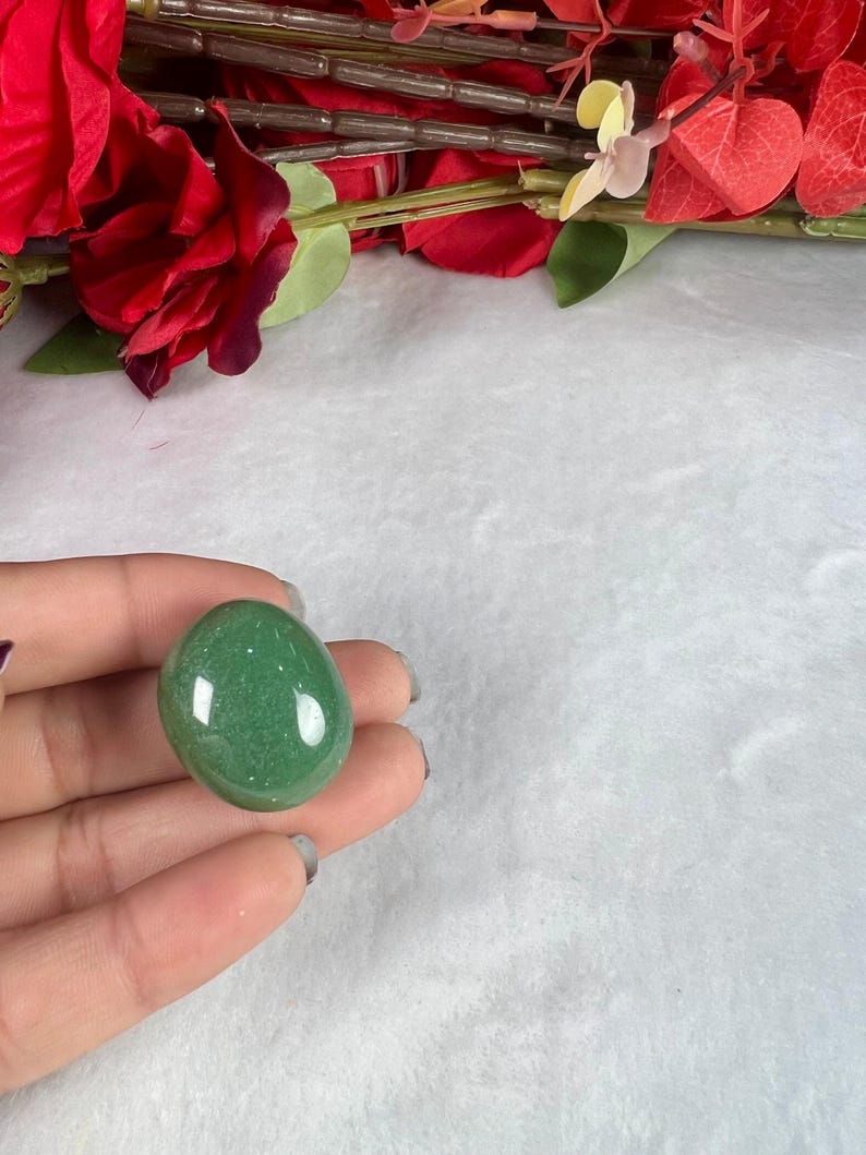 May include: A smooth, oval-shaped green gemstone, held in a hand, with a polished surface. The stone has a speckled appearance and reflects light. Red flowers and greenery are visible in the background, set against a white, textured surface.