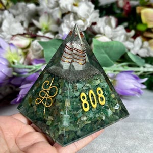 May include: A green pyramid-shaped orgone with a clear apex, containing a quartz crystal wrapped in copper wire. The base is filled with green stones and gold-colored symbols and the number "808".