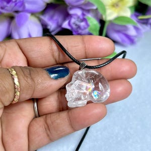 May include: Clear quartz crystal skull pendant on a black cord. The skull pendant is held in a hand, with a gold ring and a silver ring on the fingers. Purple flowers are in the background.