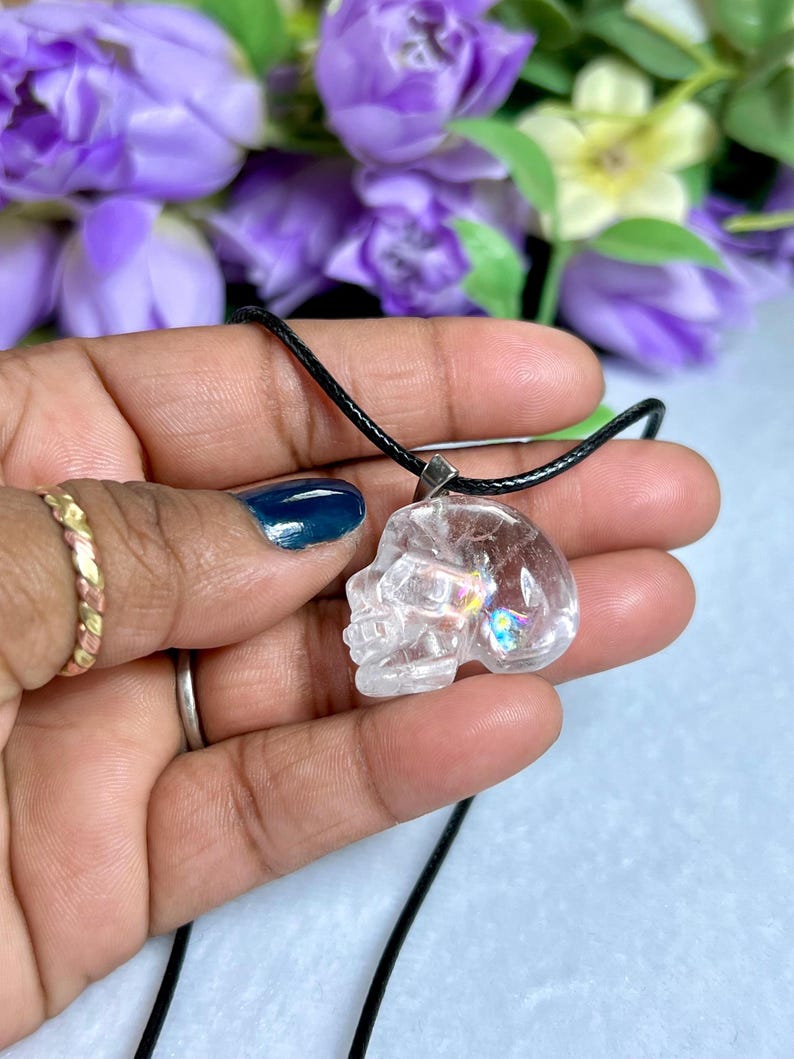 May include: Clear quartz crystal skull pendant on a black cord. The skull pendant is held in a hand, with a gold ring and a silver ring on the fingers. Purple flowers are in the background.