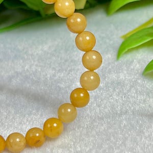 May include: A bracelet featuring round, golden-yellow beads. The beads have a smooth, polished appearance and are strung together to form a flexible band. The bracelet is set against a soft, white background with green foliage in the background.