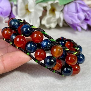 May include: A multi-strand bracelet featuring polished spherical beads in deep blue and translucent orange, woven together with dark brown cord and accented by thin, bright green thread details throughout the structure of the jewelry piece.
