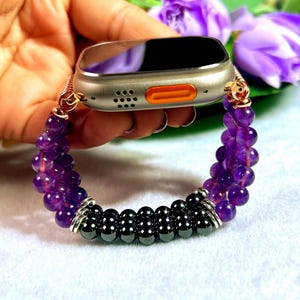 May include: A silver smartwatch with an orange button and a purple beaded band. The band features amethyst and black beads, connected with silver-toned metal. The watch is held against a background of purple flowers.