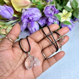 May include: A clear quartz crystal skull pendant on a black cord necklace. The skull pendant is carved with detailed features and hangs from a silver-colored bail. The necklace is displayed against a backdrop of purple flowers.